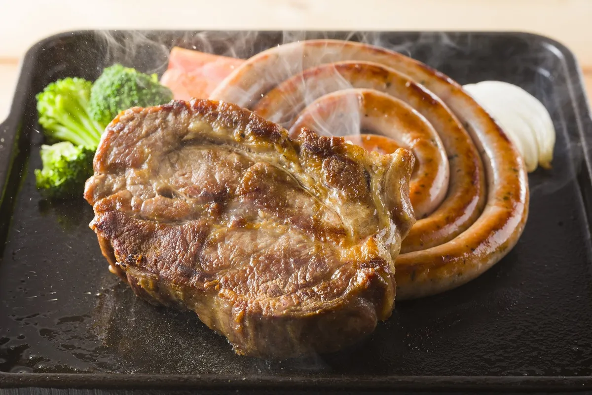 鉄板の上で湯気を立てて焼かれる、大きなステーキと渦巻き状のソーセージ